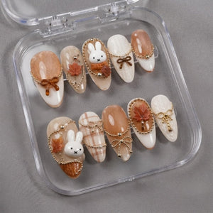 A set of ten almond-shaped press-on nails in a clear plastic case. The nails feature a color palette of white, cream, and brown, decorated with 3D white rabbit figures, brown maple leaves, gold-toned chains, and small bows.