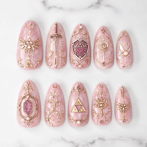 A set of ten almond-shaped press-on nails with a light pink marble base. Each nail features intricate gold-toned 3D embellishments, including symbols like the Triforce, a sword, a shield, a fairy, an eye, and floral filigree patterns.