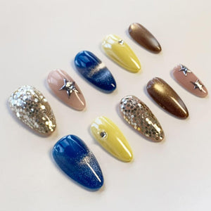 A set of ten almond-shaped press-on nails featuring various designs including solid yellow, deep blue with a glitter gradient, metallic bronze, nude with silver star accents, and full silver hexagonal glitter coverage.