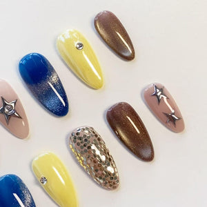 A set of almond-shaped press-on nails featuring various designs including solid yellow with a single rhinestone, deep blue with a glitter gradient, brown shimmer, nude with silver star charms, and nude with silver circular sequins.