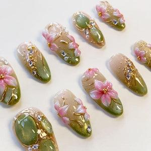 A set of ten almond-shaped press-on nails featuring a gradient from beige to olive green. Each nail is decorated with three-dimensional pink floral accents, gold metallic lines, small white pearls, and clear rhinestones.