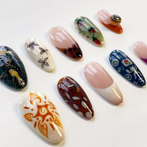 A collection of eleven almond-shaped artificial nail tips featuring diverse designs including a sun motif, floral patterns, strawberry illustrations, abstract shapes, and French tips with various colors like brown, blue, green, and white.