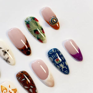 A collection of almond-shaped press-on nails featuring various designs including a strawberry pattern, a blue and white folk-art style print, a brown tortoiseshell tip, a purple French tip, and a nail with a raised blue and yellow swirl.