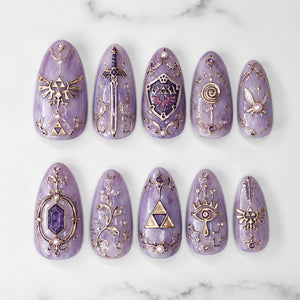 A set of ten almond-shaped press-on nails in a marbled lavender finish, featuring intricate gold-toned metallic embellishments including a sword, shield, triforce symbols, a fairy, and ornate filigree patterns with small embedded crystals.