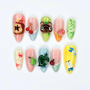 A set of ten almond-shaped press-on nails featuring Animal Crossing themed designs. The nails display various icons including a bell bag, cherries, Tom Nook, a peach, a leaf, a pear, and clothing items painted in vibrant colors.