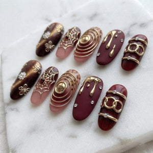 A set of ten press-on nails featuring diverse designs including dark brown cat-eye polish with floral accents, spider webs with red gems, concentric gold rings on leopard print, matte burgundy with gold drips, and gold chain links with crystals.