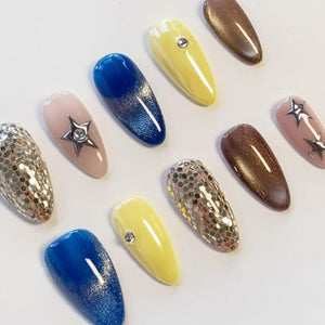 A set of ten almond-shaped press-on nails featuring various designs including solid yellow with a single rhinestone, deep blue with a glitter gradient, brown shimmer, silver hexagonal sequins, and nude bases with silver star charms.