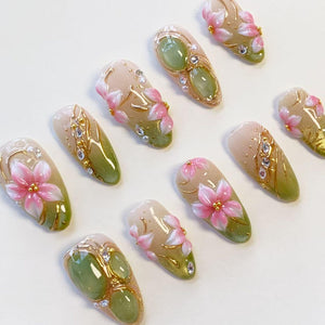 A set of ten almond-shaped press-on nails featuring a gradient design transitioning from a nude base to a sage green tip. The nails are decorated with hand-painted pink flowers, gold metallic accents, small clear rhinestones, and pearls.