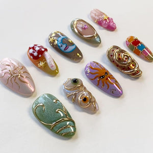 A collection of ten almond-shaped press-on nails featuring diverse designs including a gold sun, a red mushroom, a crescent moon, a purple sun face, gold swirls, and colorful geometric patterns, all accented with gold foil and 3D details.