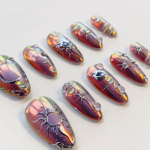 A set of ten almond-shaped press-on nails featuring a metallic, iridescent purple and red finish. Each nail is decorated with raised silver celestial motifs, including suns, stars, and a planet with rings, accented by small clear gems.