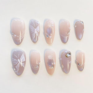 A set of ten almond-shaped press-on nails in a soft nude and mauve gradient. The design features hand-painted white butterfly wings, delicate silver chains, small white pearls, and clear rhinestones arranged on individual nails.
