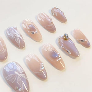 A set of almond-shaped press-on nails in a soft nude pink tone. The nails feature white hand-painted bow designs, small clear rhinestones, and tiny white pearl accents arranged in various patterns across the glossy surface.