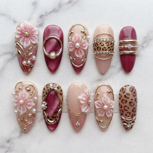 A set of ten almond-shaped press-on nails featuring a mix of designs including 3D pink flowers, gold metal accents, white pearls, leopard print patterns, and deep magenta cat-eye polish. Each nail is decorated with intricate gold filigree.