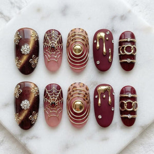 A set of ten press-on nails featuring diverse designs including gold floral accents on dark brown cat-eye polish, spider webs with red gems, leopard print with gold rings, gold liquid drips on matte burgundy, and gold bands with crystals.