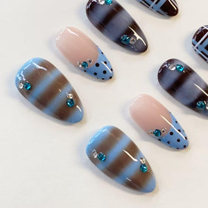 A set of almond-shaped press-on nails featuring a mix of designs. Some nails have horizontal brown and light blue stripes, while others feature a nude base with light blue tips decorated with small black polka dots and teal rhinestones.
