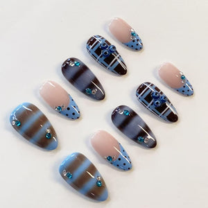A set of ten almond-shaped press-on nails featuring a variety of designs including blue and brown horizontal stripes, blue and black plaid patterns, and light pink tips with blue polka dots, all accented with small blue and clear rhinestones.