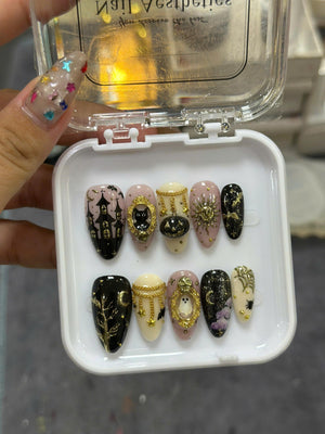 A set of ten press-on nails in a white plastic case, featuring Halloween-themed designs including a haunted house, a black cat in a gold frame, a pumpkin, a sun, a ghost, spider webs, and bats with gold and black accents.