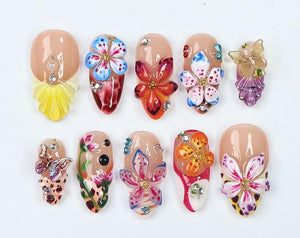 A collection of ten artificial nails, each adorned with intricate floral designs and embellishments. The nails feature a variety of colors, including pink, blue, orange, and yellow, with sparkling rhi