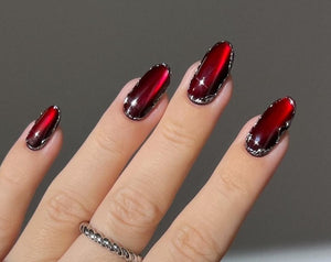 Hand with long, oval-shaped, red nails with a metallic silver border. Two silver rings are on the fingers. The nails have a glossy finish. The hand is wearing a white long-sleeved shirt.