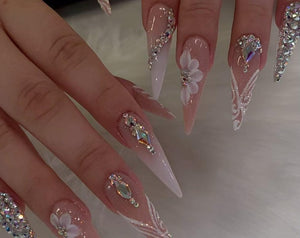 Long, pointed acrylic nails with a gradient effect, transitioning from a natural pink to white. Embellished with rhinestones, 3D white flowers, and intricate white lace designs. A glamorous and detail