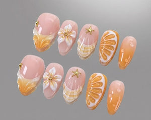A set of ten press-on nails featuring a light pink base with orange gradient tips. Designs include white 3D flowers, gold starfish charms, small gold studs, and raised orange slice patterns with white