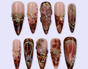A set of ten almond-shaped press-on nails featuring a nude base with brown leopard print patterns, gold metallic accents, raised floral designs, and small pearl embellishments. The nails are arranged