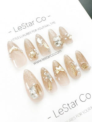 Set of ten nude-colored false nails with gold glitter and various rhinestones and pearl accents. The nails are arranged in two rows on a white surface. The text "LeStar Co - LITTLE LUXURIES FOR YOUR N