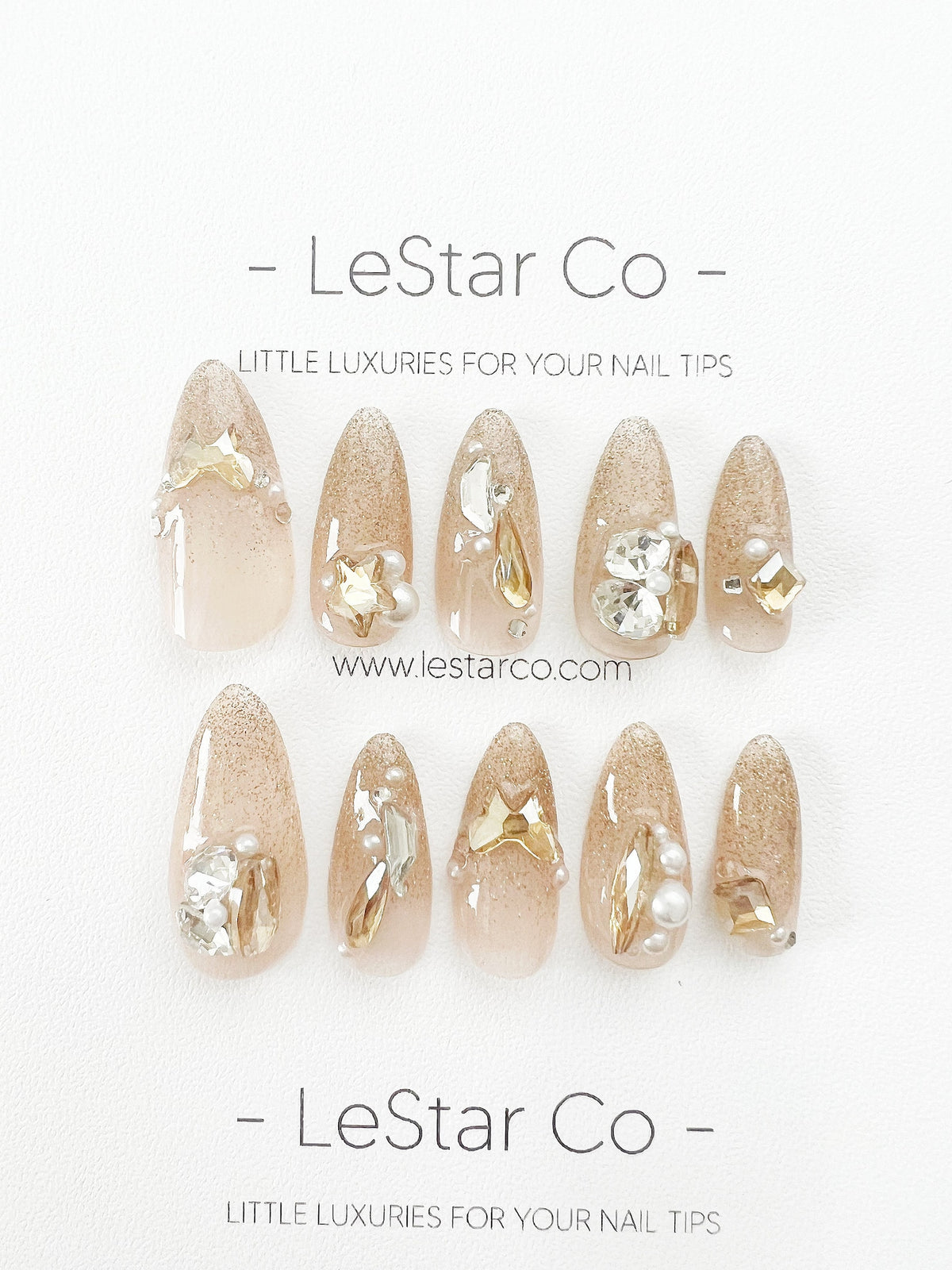Ten nude-colored, almond-shaped press-on nails with gold glitter and various embellishments, including rhinestones, pearls, and gold bows. The nails are arranged in two rows of five. The text "LeStar 