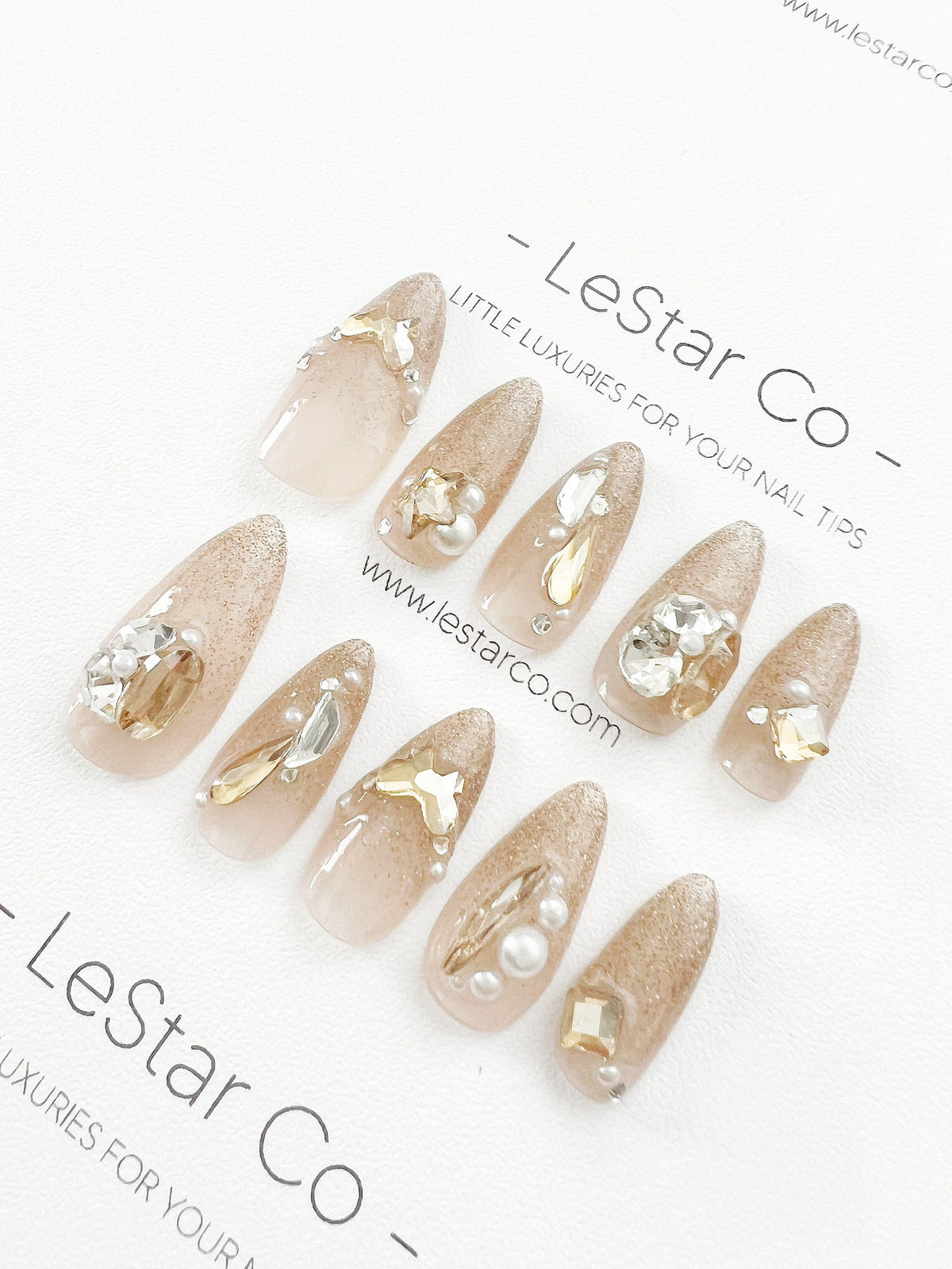 Set of ten nude-colored press-on nails with gold glitter and rhinestones. The nails are decorated with pearls and gold accents. The text "LeStar Co - Little Luxuries for Your Nail Tips" is printed on 