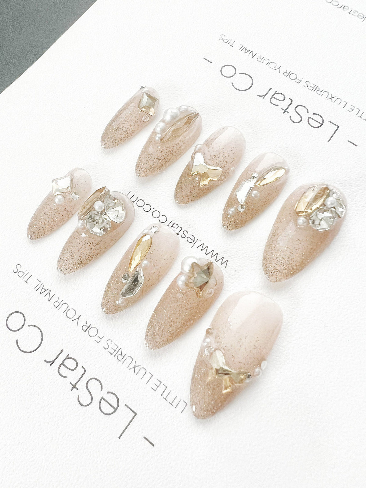 Set of ten nude-colored false nails with glitter and gold accents. Each nail is decorated with a different design featuring pearls, stars, and gold accents. The nails are arranged in a row on a white 