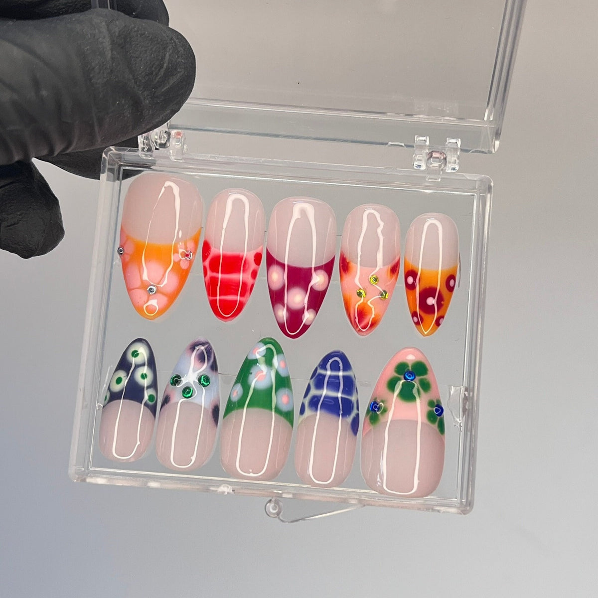 A clear plastic box with 10 oval-shaped false nails. The nails are decorated with different colorful floral patterns and rhinestones.