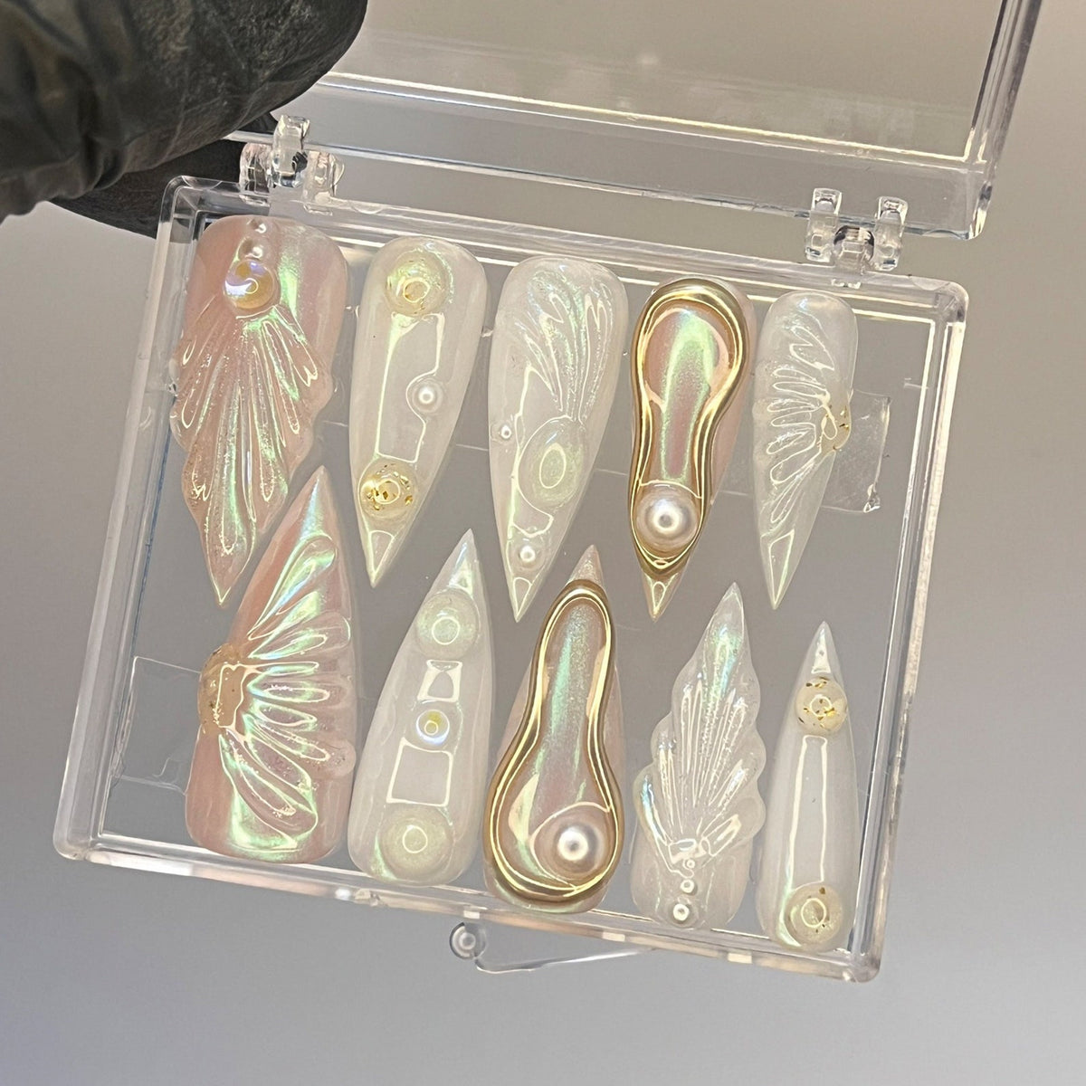 A clear plastic box with 10 iridescent nail art designs. The designs are in various shapes and sizes, including flowers, swirls, and geometric patterns. Each design has a small pearl or rhinestone in 