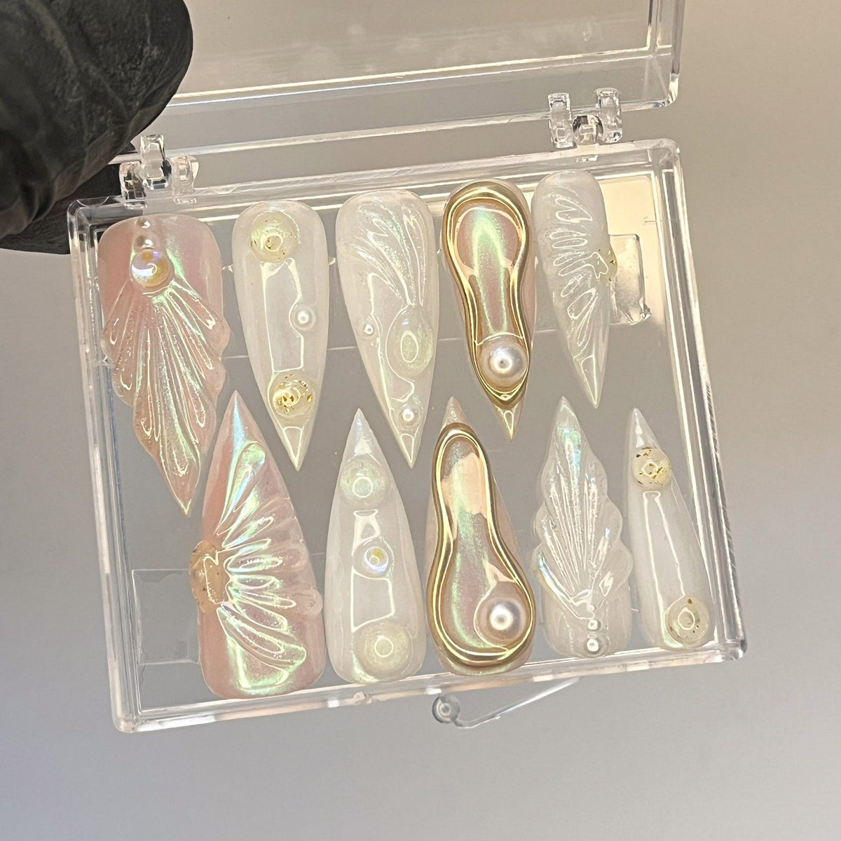 A clear plastic case with 10 iridescent nail tips. The tips have a variety of shapes and designs, including a leaf, a swirl, and a teardrop. The tips are decorated with gold accents.