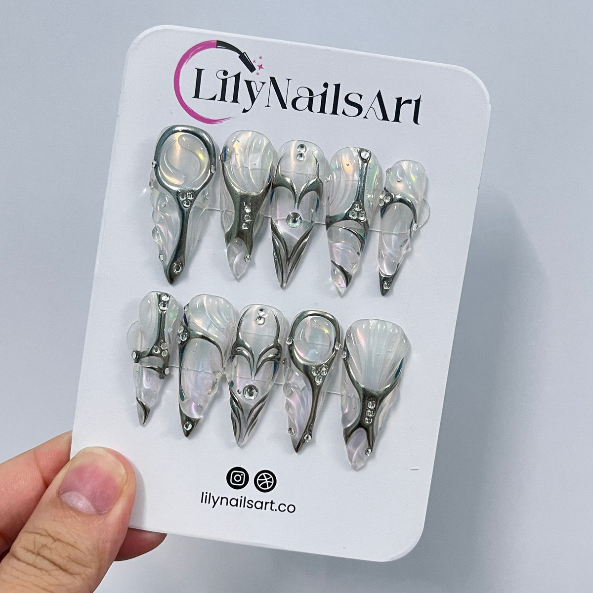 Set of ten white and silver press-on nails with a pearl-like finish. Each nail features a different design with silver accents and rhinestones. The nails are displayed on a white card with the text "L