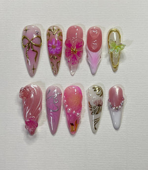 Ten pink and white long false nails with different designs. Some nails have flowers, bows, and pearls.