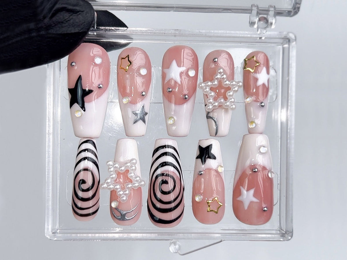 A set of ten artificial nails in a clear plastic case. The nails feature a variety of designs, including black and silver stars, white swirls, and pearl and rhinestone accents. The nails are primarily