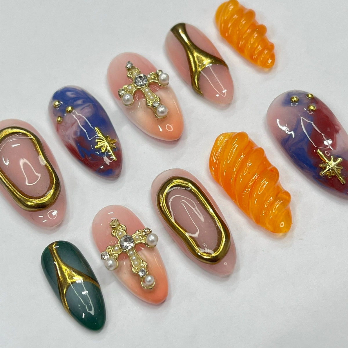 Set of ten false nails with various designs. Some nails feature a swirl pattern, others have a cross design with rhinestones, and some have a blue and white marbled design.
