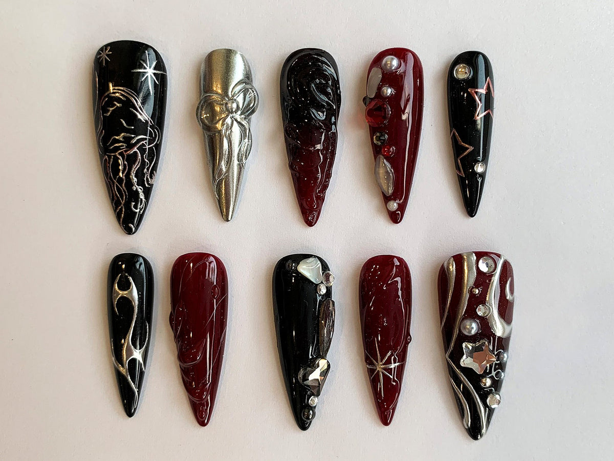 Ten long, almond-shaped false nails with different designs. Some nails are black with silver accents, others are red with silver accents, and one is silver with a bow design.