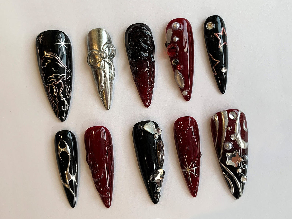 Ten long, almond-shaped false nails with different designs. Some nails are black with silver accents, others are red with gold accents, and one is silver with a bow design.