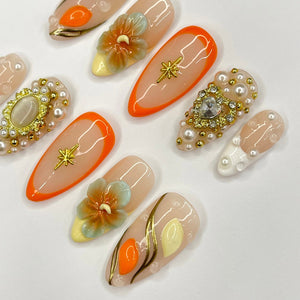 Set of ten peach-colored fake nails with various designs. Some nails have gold accents, pearl embellishments, and 3D floral designs.