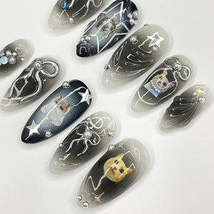 Close-up of ten long, oval-shaped, white and black nail tips with a gradient design. Each nail tip features a different design, including stars, bows, and a cat illustration. Some nail tips have silver glitter accents.