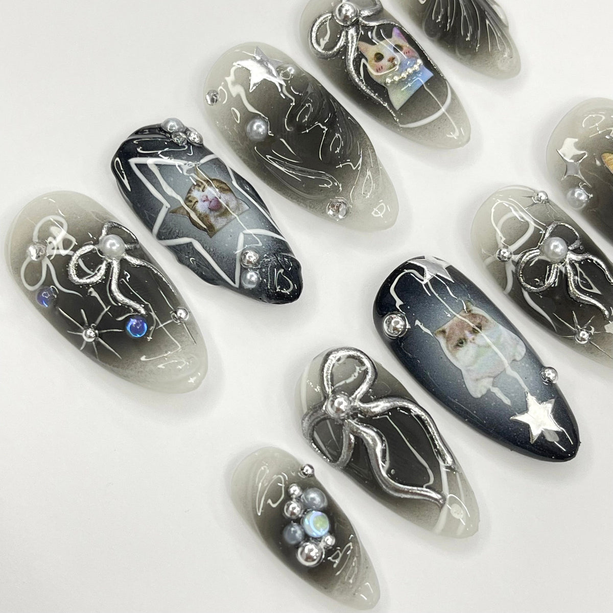 Set of ten black and white oval-shaped false nails with a gradient design. Each nail features a different design, including a cat, a star, and a bow. Some nails have silver glitter and pearls.