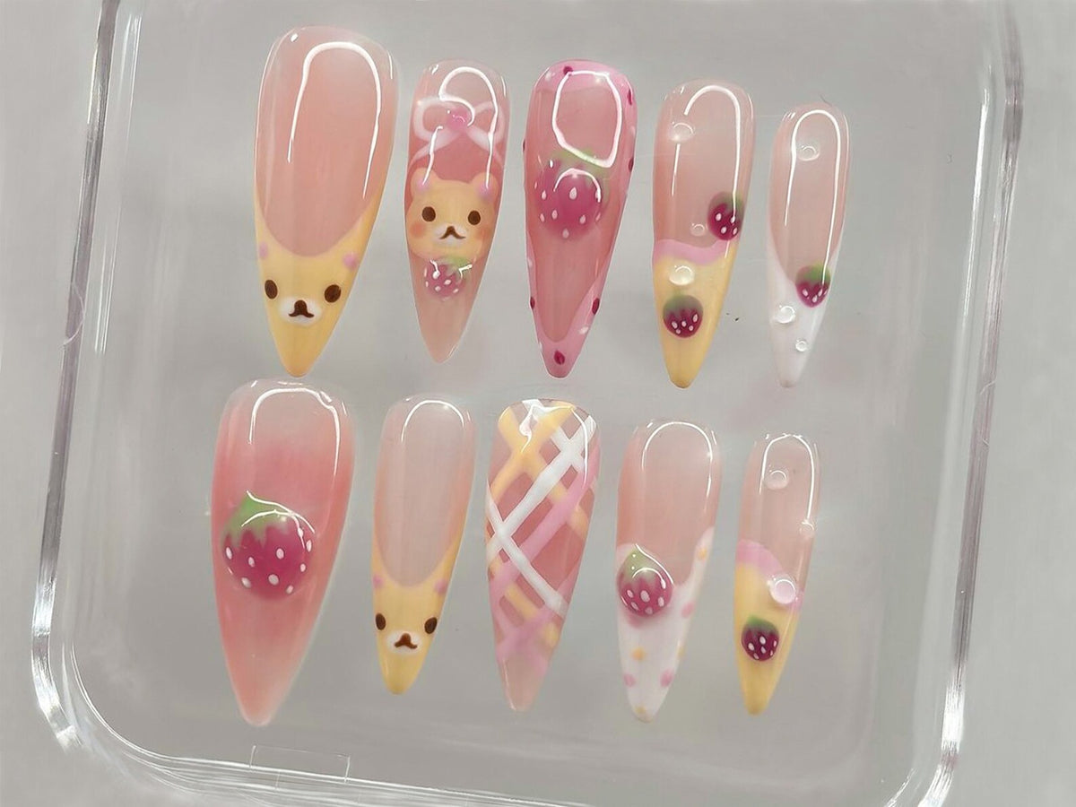 A set of ten artificial nails with a variety of designs. The nails feature a light pink base with designs including strawberries, a bear, and plaid patterns. Some nails have a yellow tip.