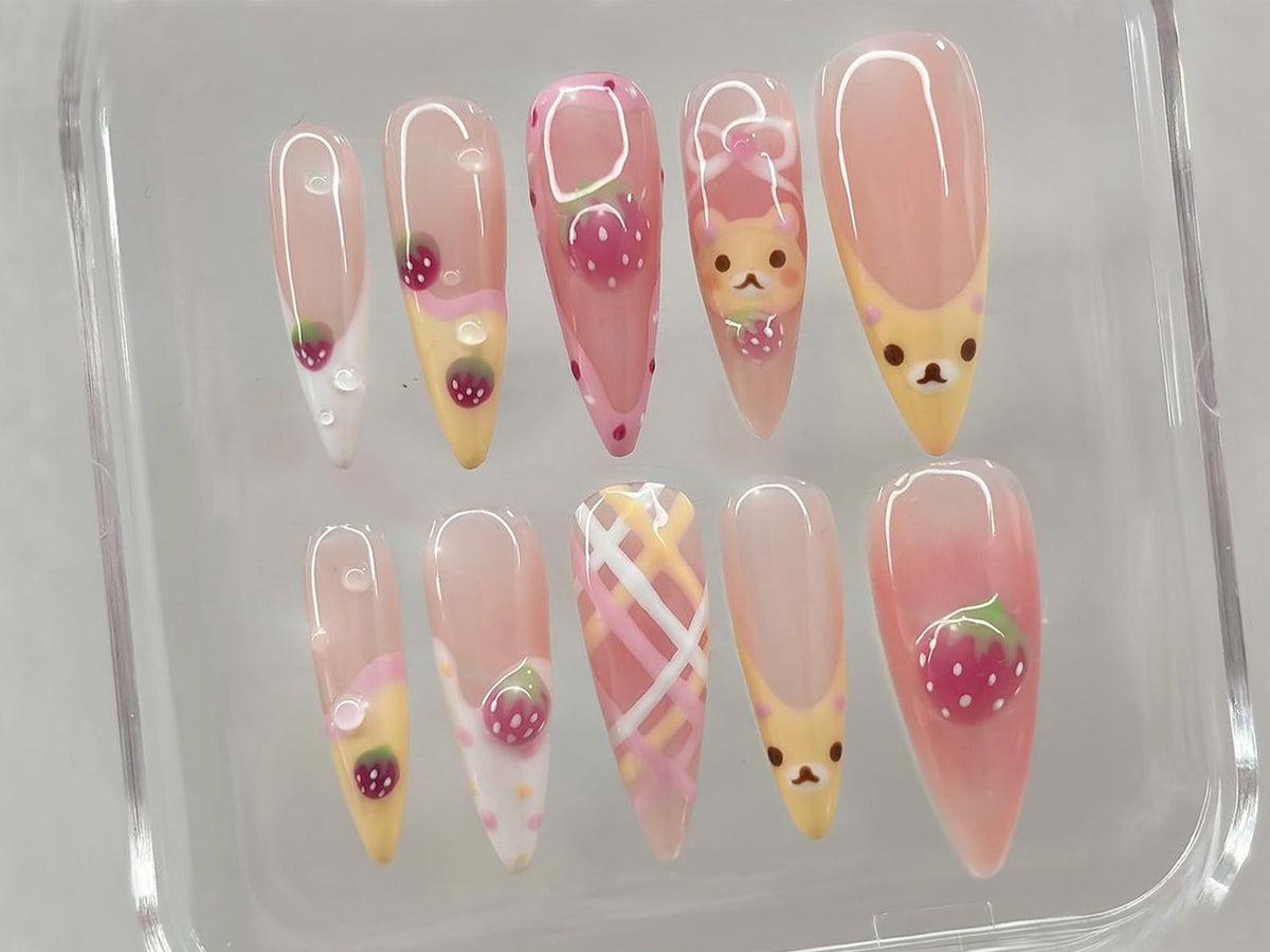 A set of ten artificial nails with a variety of designs. The nails feature a light pink base with strawberry, bear, and plaid patterns. Some nails have a clear, glossy finish with small water droplet 