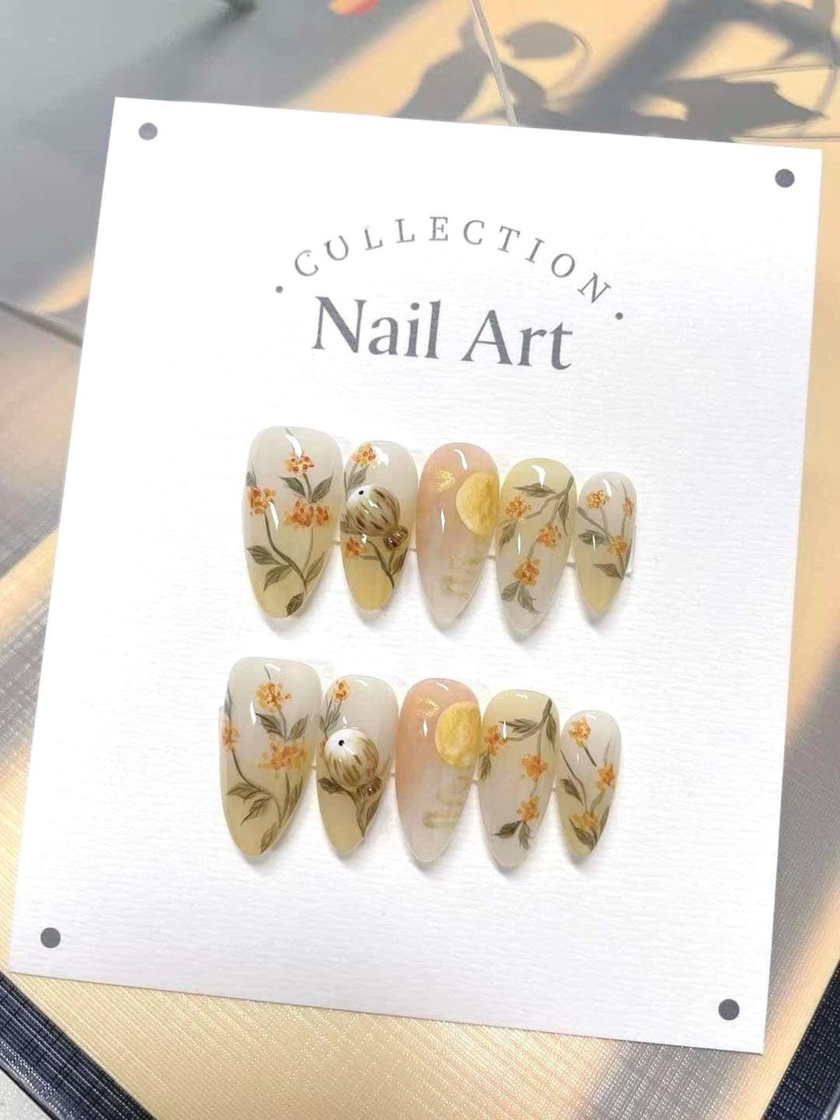 A white card with the text "COLLECTION. Nail Art." printed on it. The card displays ten artificial nails with a floral and moon design. The nails are painted in a light beige color with yellow flowers