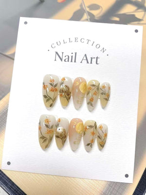 A white card with the text "COLLECTION. Nail Art." printed on it. The card displays ten artificial nails with a floral and moon design. The nails are painted in a light beige color with yellow flowers
