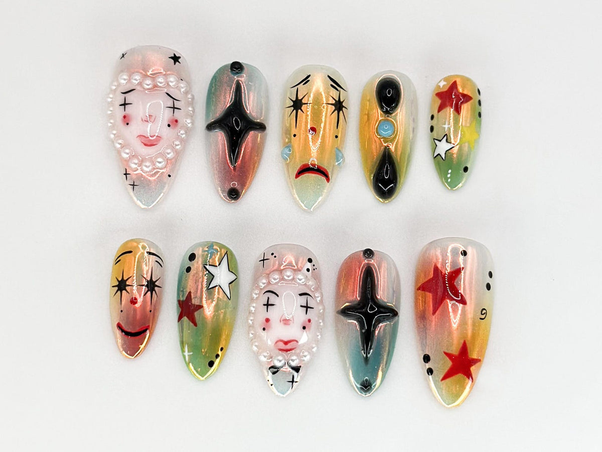 Ten false fingernails with different designs. Each nail is painted with a different design, including a face with a pearl necklace, a black star, a crying face, a black circle with a turquoise gem, a 
