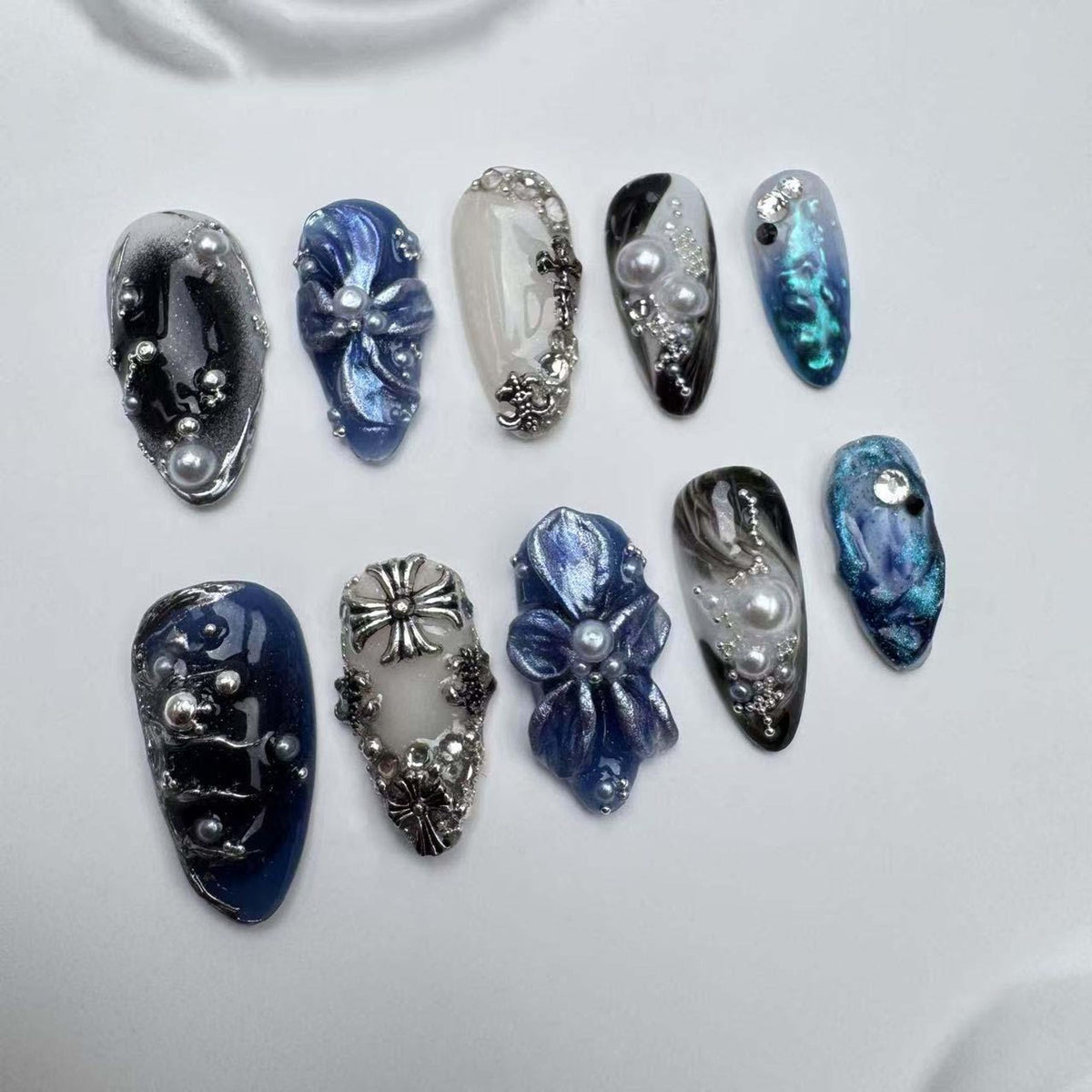 Ten blue and white nail tips with various designs, including a cross, a flower, and a swirl pattern. Each nail tip is decorated with pearls and rhinestones.