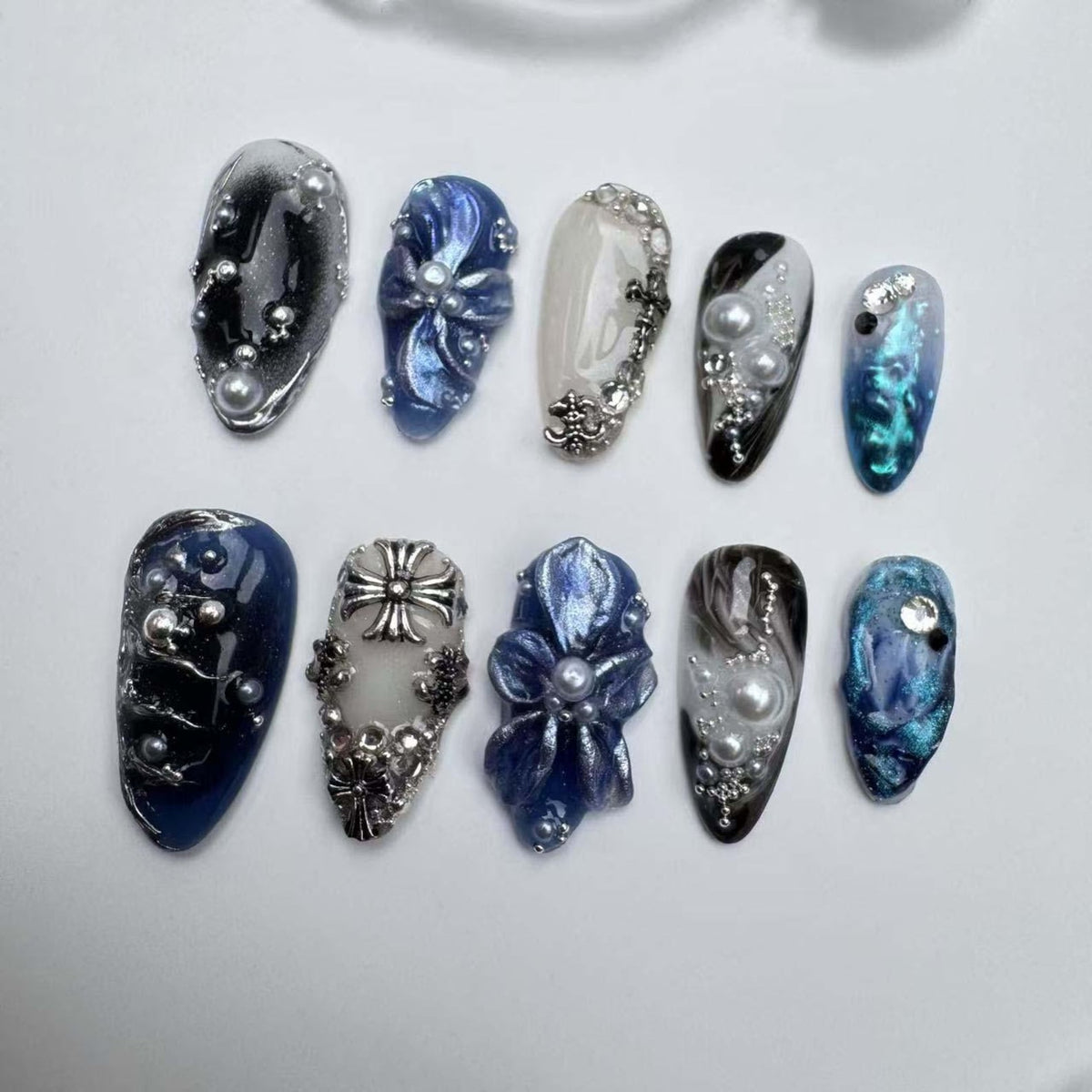 Ten blue and white nail tips with various designs, including flowers, swirls, and a cross. Some tips have pearl accents.