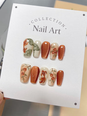 A set of ten press-on nails with a fall-themed design. The nails are a mix of white with orange and green floral designs and a shimmery copper color. The nails are displayed on a white card with the t